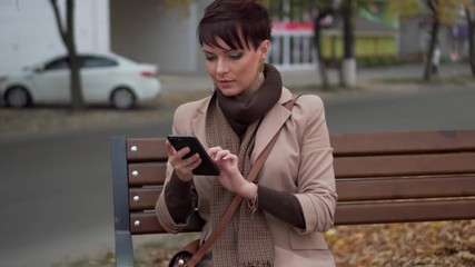 young female uses smartphone while sitting on bench against background of city