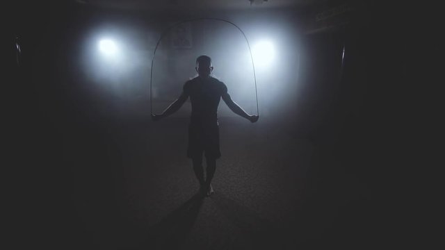 Boxer does some jump rope exercises in dark room under light. Fighter training in smoky studio in slow motion. Silhouette on dark background
