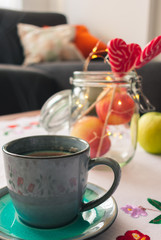 Close up of a blue cup of coffee on an embroided linen, apples in a jar and heart shaped lollipops with lights on a wooden table. Autumn or holiday interior decoration ideas.