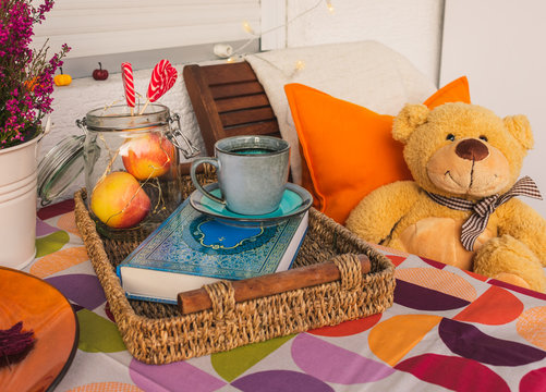 View of a teddy bear sitting in a balcony chair in autumn, composition with coffee, book, apples in a jar, light chain and heart shaped lollipops. Autumn colors and balcony decoration