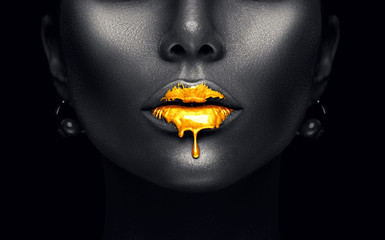 Gold paint drips from the sexy lips, golden liquid drops on beautiful model girl's mouth, creative abstract makeup. Beauty woman face isolated on black - obrazy, fototapety, plakaty