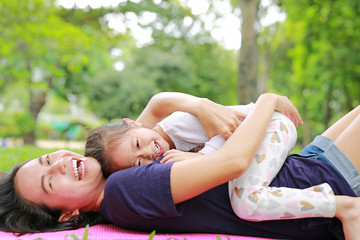 Obraz na płótnie Canvas Happy Asian mom embrace her daughter lying in the green garden with looking camera. Funny mother and child girl playing in summer park.