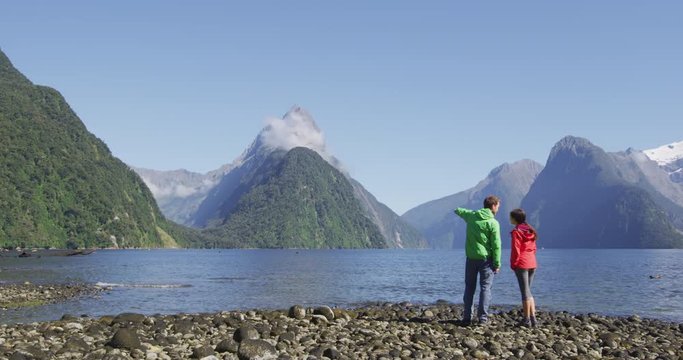 Milford Sound tourist couple hiking in New Zealand enjoying iconic view of Mitre Peak, Fjord and famous tourist destination in Fiordland National Park,.