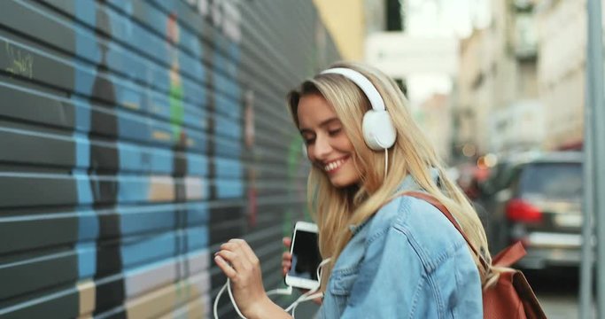 Portrait shot of the happy pretty young woman with a blond hair having fun and dancing a little while listening to the music on the smartphone player and with headphones. Outdoors.