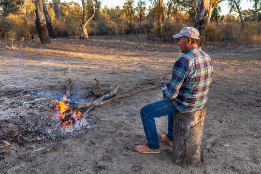 Mature man sitting on stump and enjoying a beer as he contemplates the World while looking into a campfire in the Australian outback.