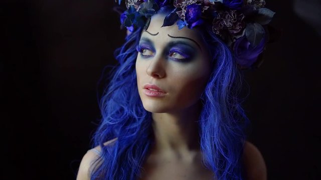 Fashion. Portrait of a young woman with frightening makeup on Halloween slow motion. Painted face of a sad skull with blue eyelashes and hair . Calm face. Dark background.