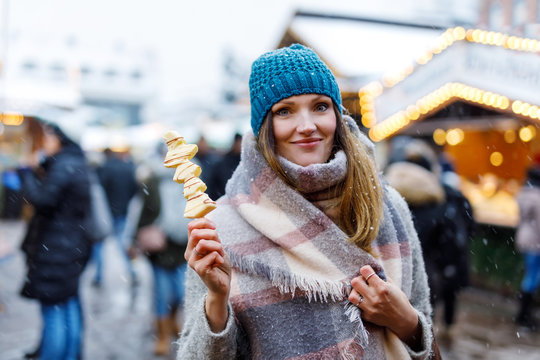 Beautiful young woman eating white chocolate covered fruits on skewer on traditional German Christmas market. Happy girl on traditional family market in Germany, Munich during snowy day.