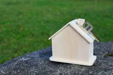 Small wooden house model with key on grey stone on green blurred grass background with copy space for text. House building, loan, real estate or buying or renting new home concept.