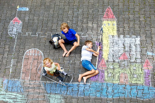 Two little kids boys and cute toddler girl drawing knight castle with colorful chalks on asphalt. Happy siblings and friends having fun with creating chalk picture and painting