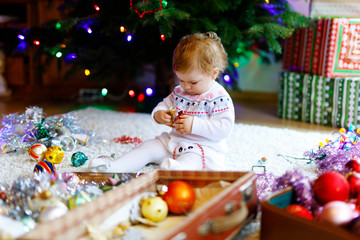 Fototapeta na wymiar Adorable baby girl holding colorful vintage xmas toy and ball in cute hands. Little child in festive clothes decorating Christmas tree with family. First celebration of traditional feast