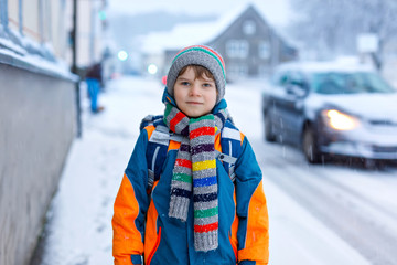 Little school kid boy of elementary class walking to school during snowfall. Early morning and snowy streets in city. Student with backpack or satchel in colorful winter clothes.