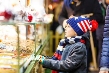 Little cute kid boy near sweet stand with gingerbread and nuts. Happy child on Christmas market in Germany. Traditional leisure for families on xmas. Holiday, celebration, tradition, childhood