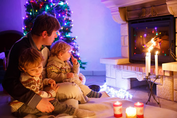 Father and two little toddler boys sitting by chimney, candles and fireplace and looking on fire. Family celebrating Christmas. With Xmas tree and lights on background. Kids happy about gifts..
