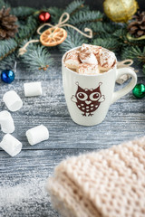 Mug with hot chocolate and marshmallow on christmas background with fir branches, cones and balls on grey wooden table.