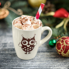 Mug with hot chocolate and marshmallow on christmas background with fir branches, cones and balls on grey wooden table.