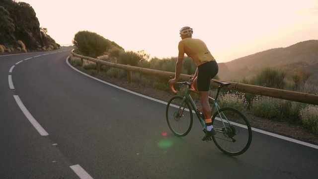 A professional cyclist in a helmet and sports equipment rides on a mountain serpentine at sunset in the direction of the volcano. Steadicam