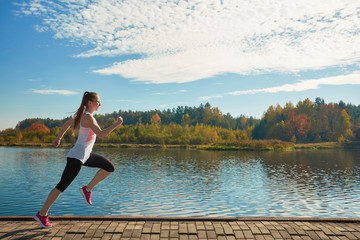 Athletic young redhead white smiling woman in pink sneakers jogging along river on the riverfront....