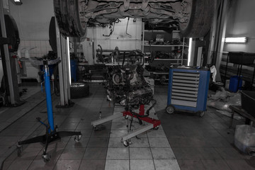 A sports car raised on a lift for repair and under it a detached engine suspended on a blue crane and a gear box on a lifting table in a vehicle repair shop with wheels lying on the sides