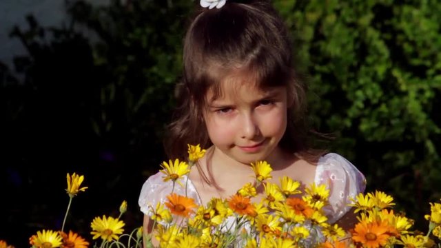 A young girl sits by bunches of yellow and orange African Daisies and waves.  She is obviously allergic to them and is squinting, blinking, and rubbing her irritated eyes.