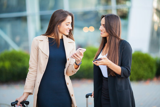 Image of two joyous european women looking at smartphone, while standing with luggage near airport waiting for flight or after departure. Air travel concept