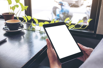 Mockup image of a woman holding black tablet pc with blank white desktop screen with green leaves and coffee on wooden table