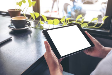 Mockup image of a woman holding black tablet pc with blank white desktop screen with green leaves and coffee on wooden table