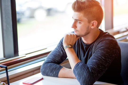 Young handsome bearded caucasian man sitting near the window, looking with pensive expression at the window at a seaside restaurant. Waiting for his coffee.