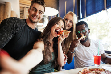 Cheerful happy multiracial friends taking selfie in pizzeria while celebrating vacation time start.