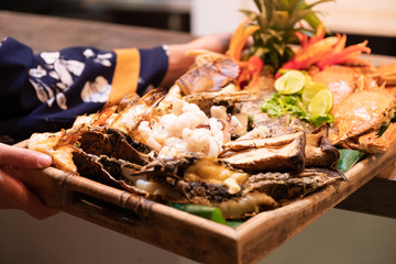 Seafood grilled (crab, fish, shrimp and shell bbg) on a wooden plate served by woman with bohemian dress costume.