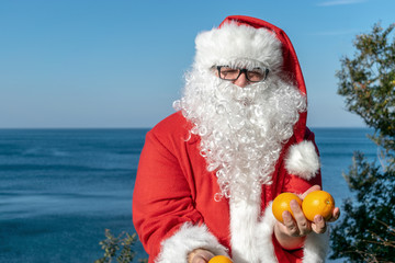 Fat man in glasses dressed as Santa holds tangerines on the ocean. Vacation and healthy lifestyle