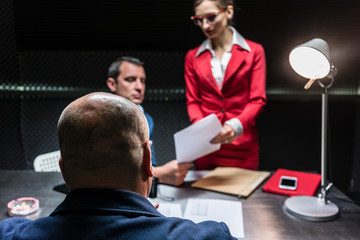 Rear view of a middle-aged suspect or witness sitting at desk during the police interrogation in front of the prosecutor and a female lawyer - obrazy, fototapety, plakaty