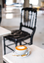 Coffee cup of Cafe' latte with heart latte art on top over the white marble table and blurred black chair in background