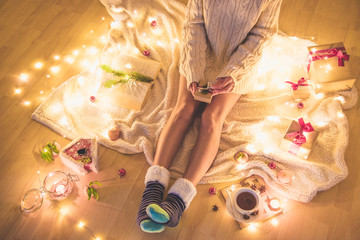 Top view of girl (young woman) sitting on the blanket laid on the floor with open book and cup of tea opening small Christmas presents - winter holidays (new year, Xmas) celebration concept