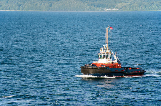 A tugboat with a Danish flag navigates on the sea, in the background the wooded coast can be seen
