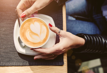 Female hands with red manicure holding cup of cappuccino on table in cafe.