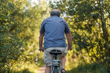 Mature man is riding bicycle in forest path with the bike to the far end