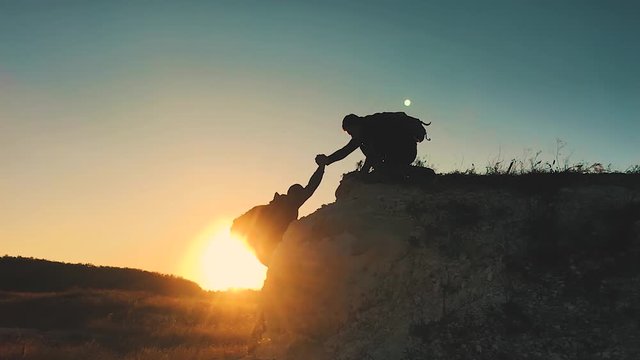 Silhouette of helping hand between two climber. two hikers on top of the mountain, a man helps a man to climb a sheer stone. couple hiking help each other silhouette in mountains with sunlight.