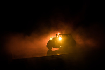 Silhouette of old vintage car in dark foggy toned background with glowing lights in low light.