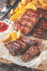 Delicious barbecued ribs on an rustic wooden chopping board with potato wedges and sauce