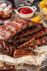 Delicious barbecued ribs on an rustic wooden chopping board with potato wedges and sauce, glass of drink with ice