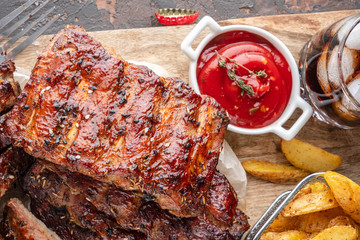 Delicious barbecued ribs on an rustic wooden chopping board with potato wedges and sauce, glass of drink with ice, top view