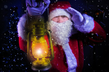 Young santa clause, carrying lantern looks through blizard of snow with christmas tree and street lamp in background