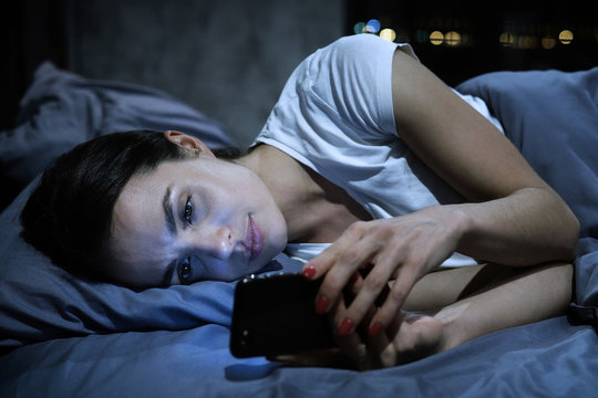 Smartphone addiction. Young tired female looking at her mobile phone screen, lying in bed late at night, scrolling her social media news feed