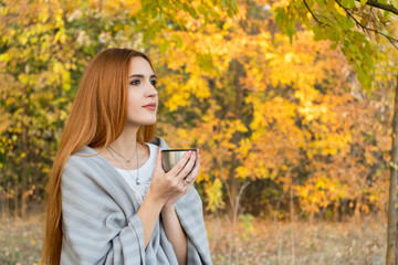 Red haired girl in the autumn forest drink tea with plaid on the shoulder