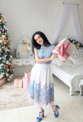 Beautiful young girl in an evening dress holds a gift in a beautiful package, a Christmas tree on the background