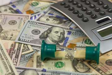 Fototapeta premium Hourglass is on dollar banknotes and calculator lies on a money background. Business and finance concept.
