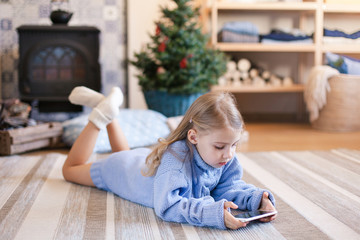 Child is using mobile phone under Christmas tree at home. Kid is playing in children games, looking at devices. Girl in blue knitted sweater with her present or gift is laying on warm floor.