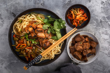 Chinese noodles with mushrooms, fried meat in a pan, vegetables and herbs are on the gray table. Top view