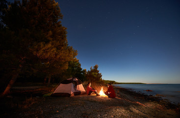 Night camping on sea shore. Happy young man and woman hikers relaxing in front of tent at campfire under evening sky on clear blue water and green forest background. Active lifestyle concept. - obrazy, fototapety, plakaty