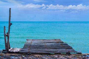 The old and dark wooden jetty in fishery village with tranquil sea and white cloudy on blue sky background.